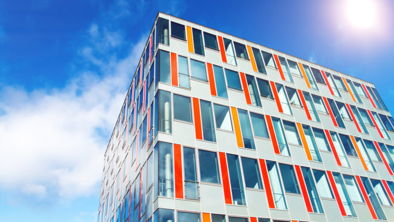 Modernes Bürogebäude mit bunter Glasfassade und spiegelnden Glasfenstern vor strahlend, blauem Himmel